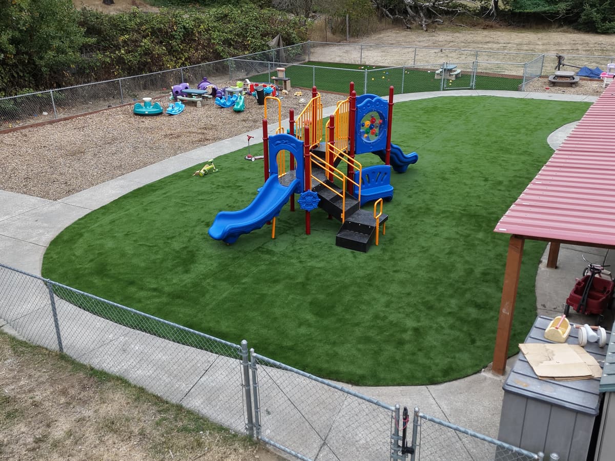The new turf play yard at Fort Nugent with the blue and yellow play structure, the bark area with ride-on toys and the covered patio to one side